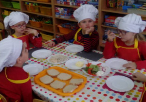 Pięcioro dzieci w strojach kucharzy siedzi przy stoliku ,na którym widać kanapki z pastą